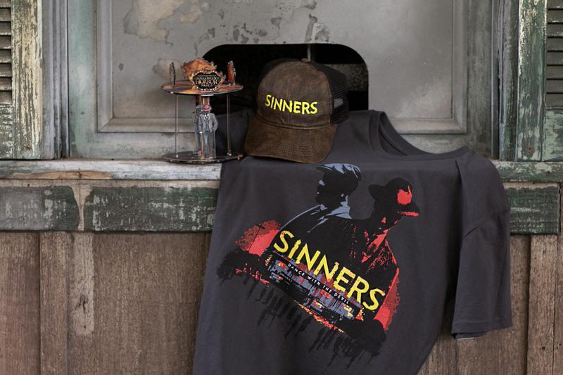 A hat and t-shirt with 'Sinners' text and promotional images and a Halloween Horror Nights commemorative carousel with a figure of Smoke and Stack in it.