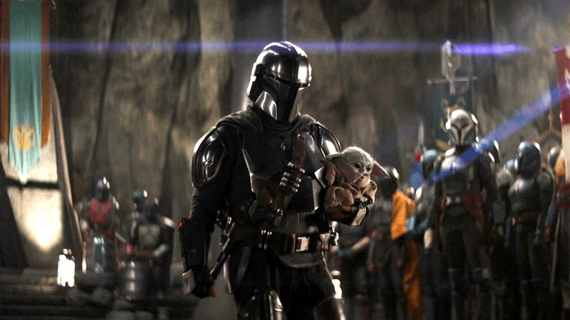 Din Djarin, in his Mandalorian armor, cradles Grogu in his arms with other Mandalorians in the background.