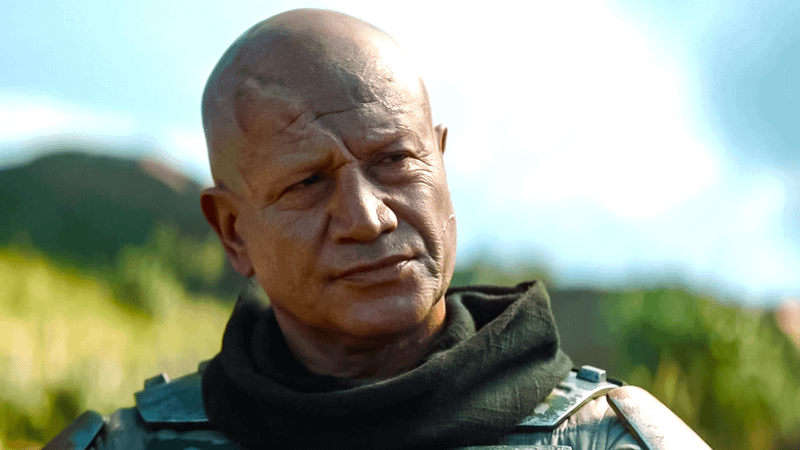 An unmasked Boba Fett stands before a lush, green landscape.