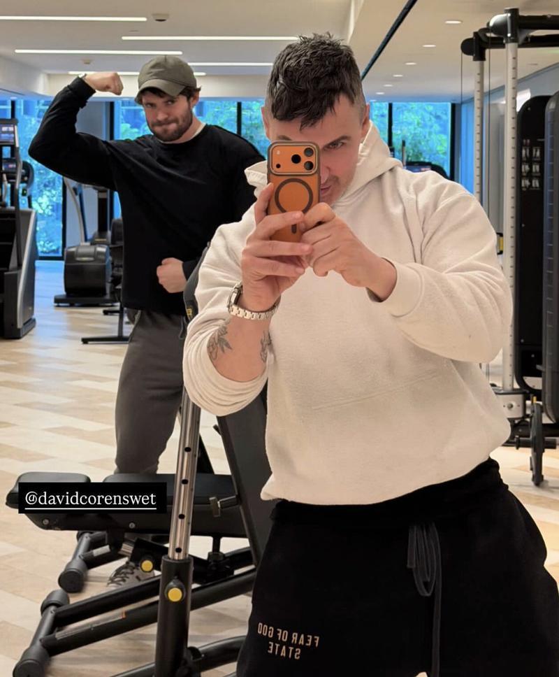 Superman 2 star David Corenswet, wearing a gray baseball cap, black sweatshirt, and gray pants and sneakers, flexes for the camera in the gym behind his trainer, Paolo Mascitti