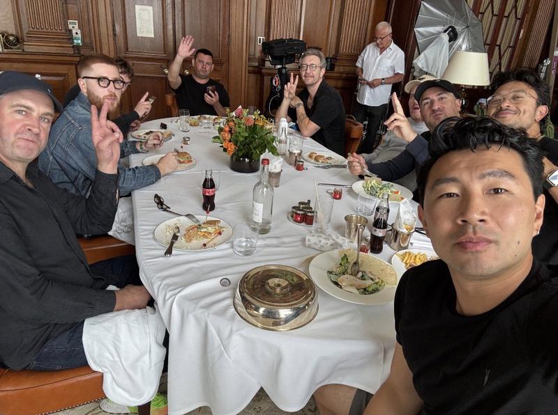 Simu Liu sitting around a table eating a dinner in a wood-panelled room