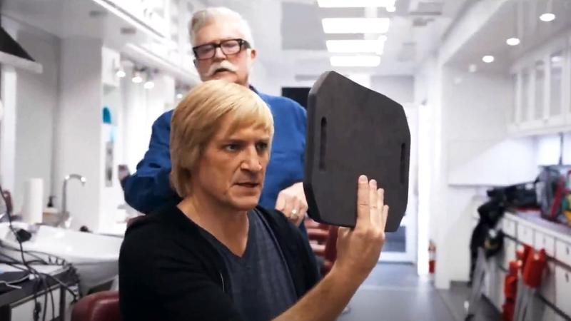 Nathan Fillion with a blond mullet holding a mirror up in Superman
