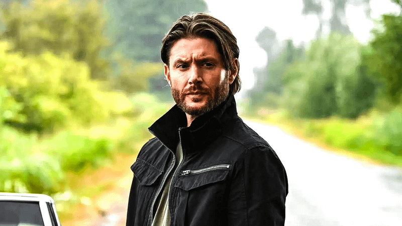 Jensen Ackles as Dean Winchester standing on a forest road in Supernatural.