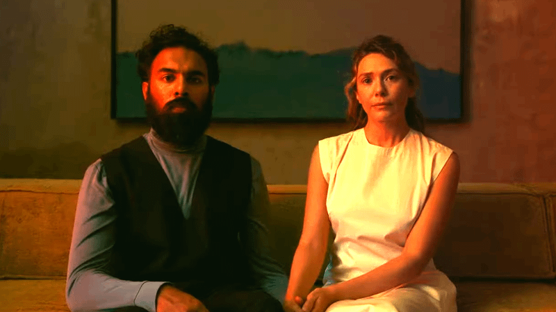 Elizabeth Olsen as Mia sitting next to Himmesh Patel as Aaryan on a couch bathed in red light in The Assessment