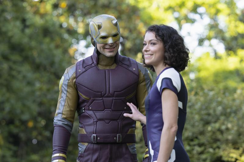 Daredevil in his yellow-and-red suit next to Jennifer Walters during the day. Green trees are behind them.