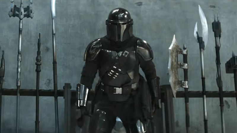 Din Djarin standing in front of an array of weapons.