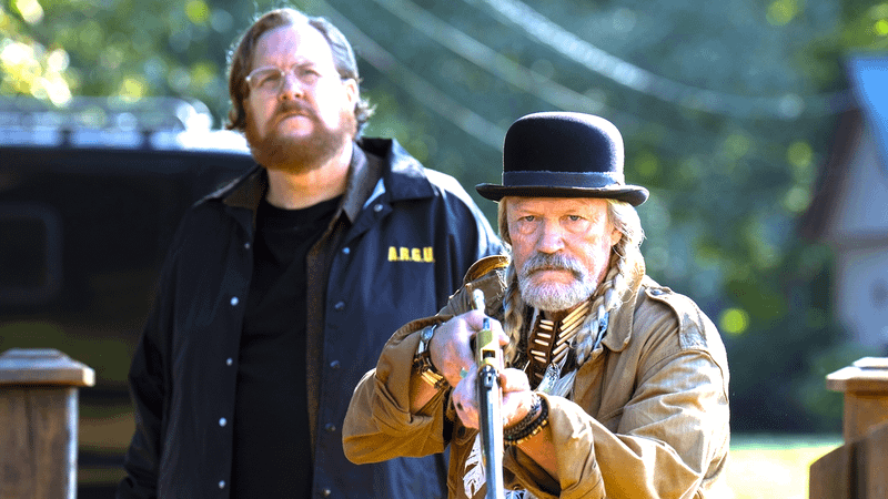 Steve Agee as John Economos looking to the sky while wearing an ARGUS jacket standing next to Michael Rooker as Red St. Wild holding up a rifle in Peacemaker