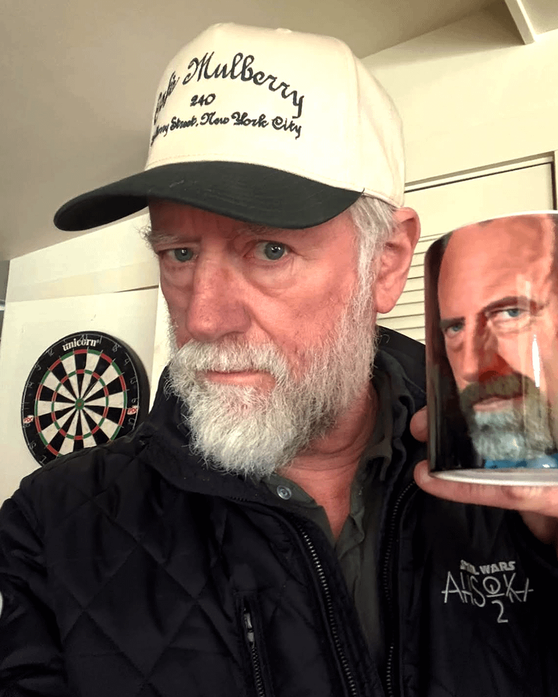 Xander Berkeley wearing a white ballcap and holding a mug up close to his face.