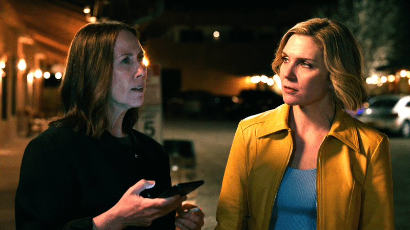 Rhea Seehorn as Carol in a yellow jacket walking next to Miriam Shor as Helen holding out her phone in Pluribus 