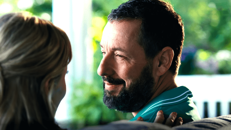 Adam Sandler in a green collared shirt sitting on a bench looking at Julie Bowen on a porch in front of greenery in a still image from Happy Gilmore 2