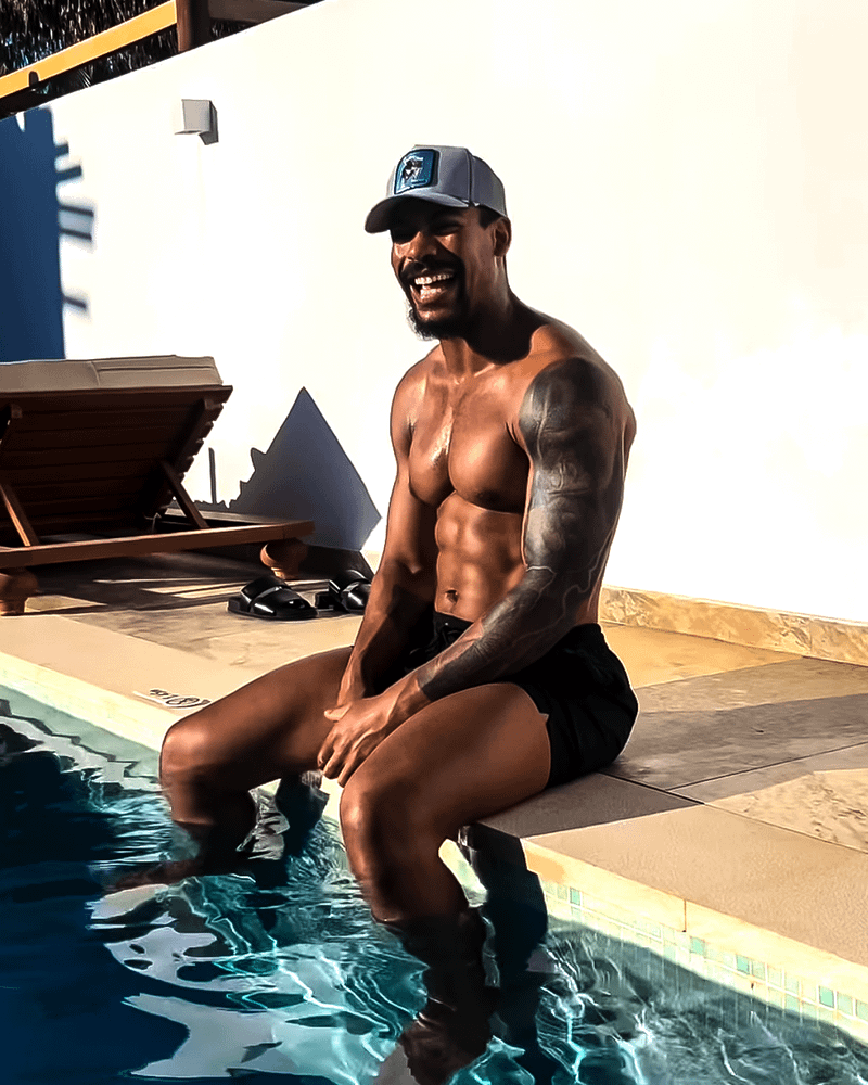 Aaron Pierre sitting by a pool in swim trunks and laughing.