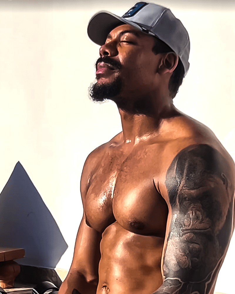 Aaron Pierre sitting shirtless with a cap on.