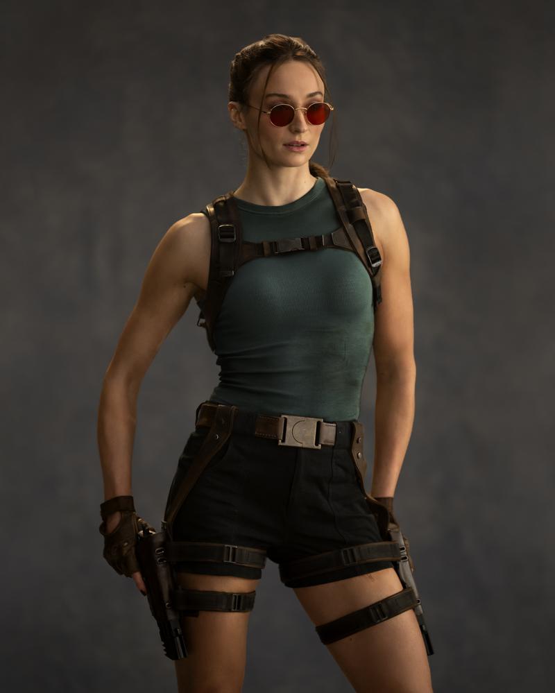 Sophie Turner poses as Lara Croft in Prime Video's upcoming Tomb Raider series, wearing a dark green tank top, black shorts, hip holsters containing dual pistols, red sunglasses, and reddish brown hair pulled away from her face. 